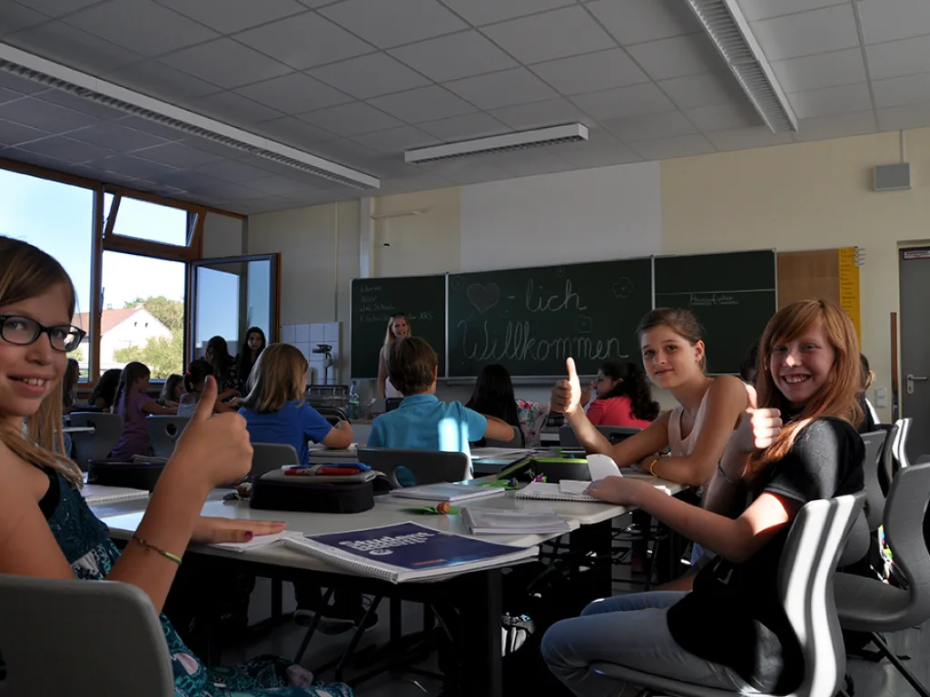 Mädchen zeigen am Tisch den Daumen nach oben, dahinter die Willkommens-Tafel.